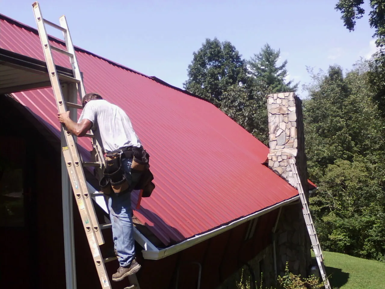 Roofer on a ladder beside a red metal roof and stone chimney — Soffit & Fascia Repair by Roofing Brothers in Pomfret